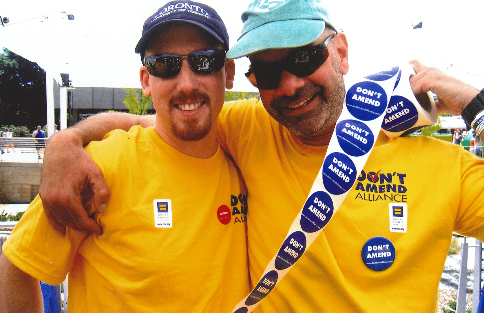 Two hugging smiling men in yellow shirts with stickers: don't amend