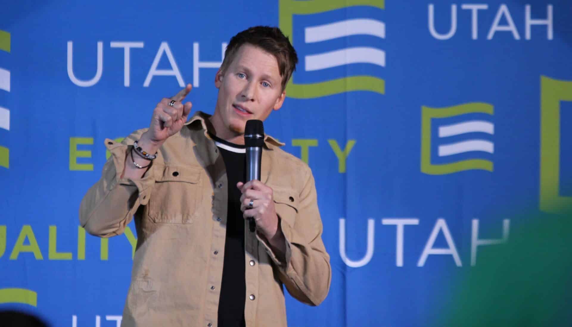 Man standing on stage in front of a blue backdrop with Equality Utah Logos, holds a microphone and points at audience.