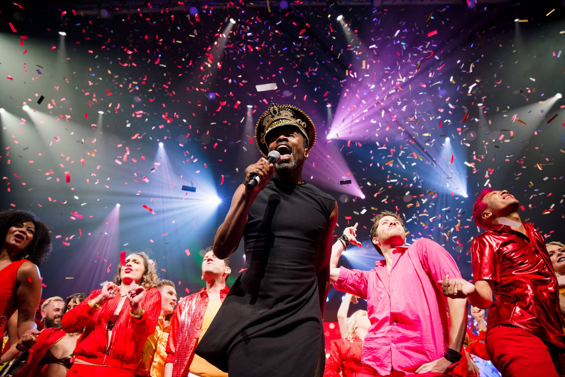 Performer sings into a microphone, surrounded by dancers dressed in red with confetti raining down