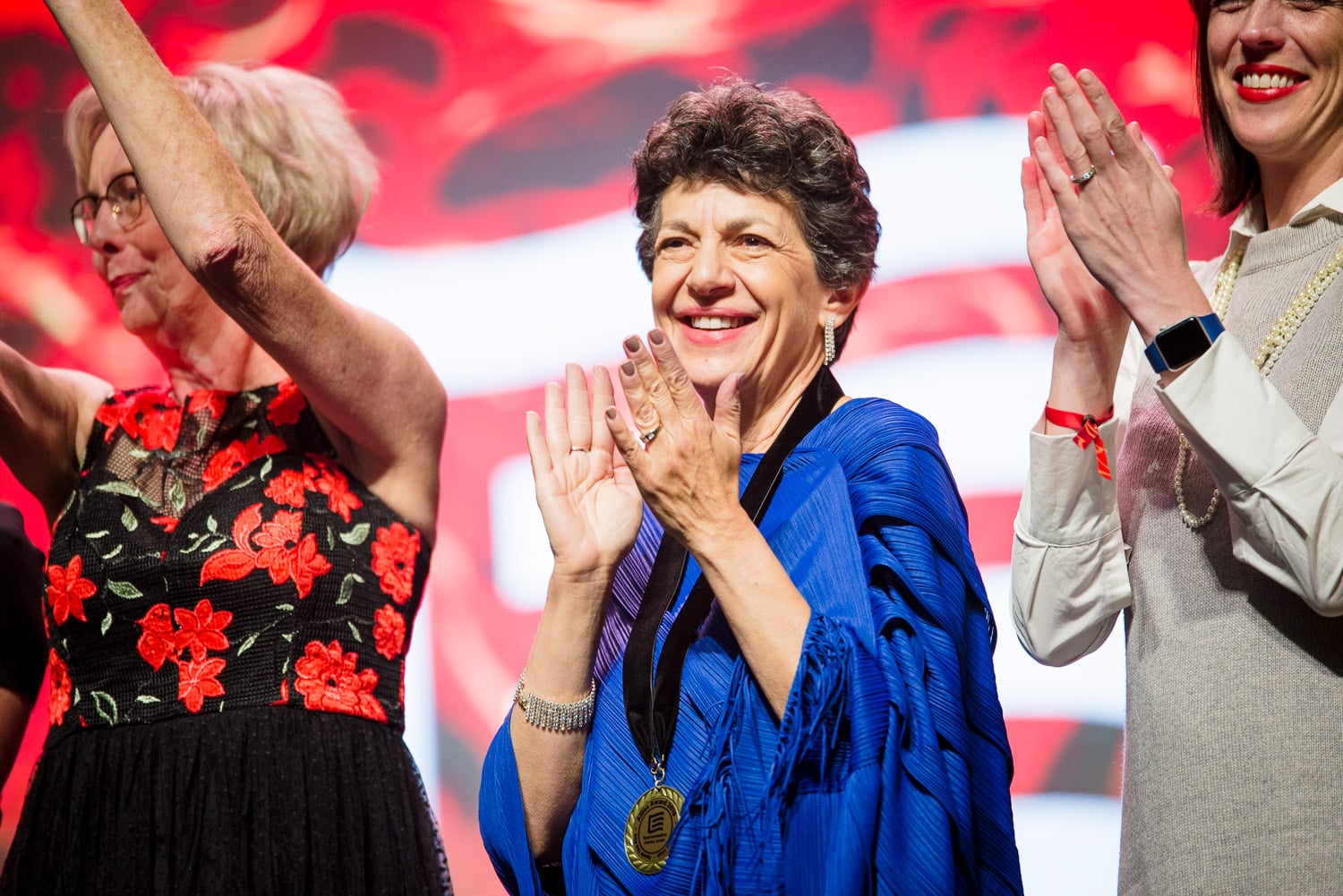 An older woman in a blue dress claps on stage, two other woman can be seen slightly out of frame