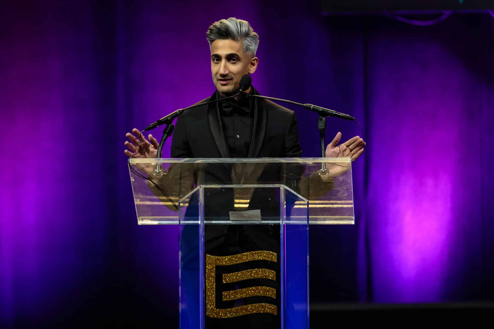 Tan France stands behind a speaker podium on a stage with a purple backdrop.