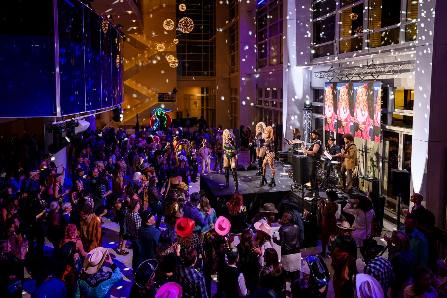 A large crowd surrounds a small stage with five drag performers.