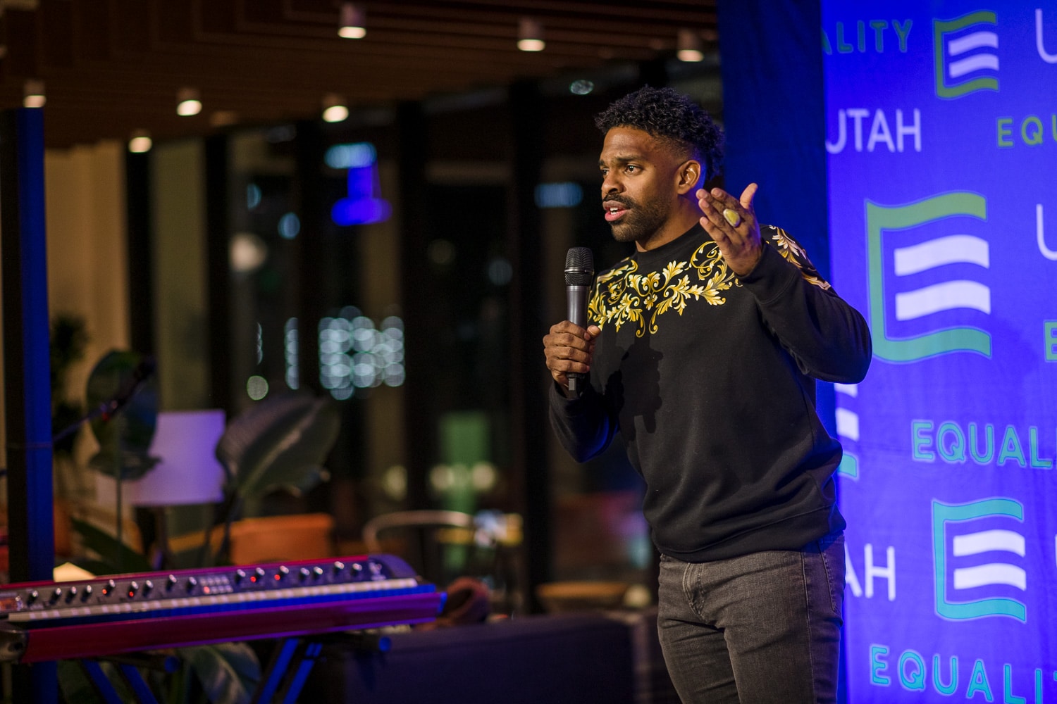 A man, wearing a black sweater with gold accents is on a stage holding a microphone speaking to an audience. The stage has a blue backdrop with 'Equality Utah' logos