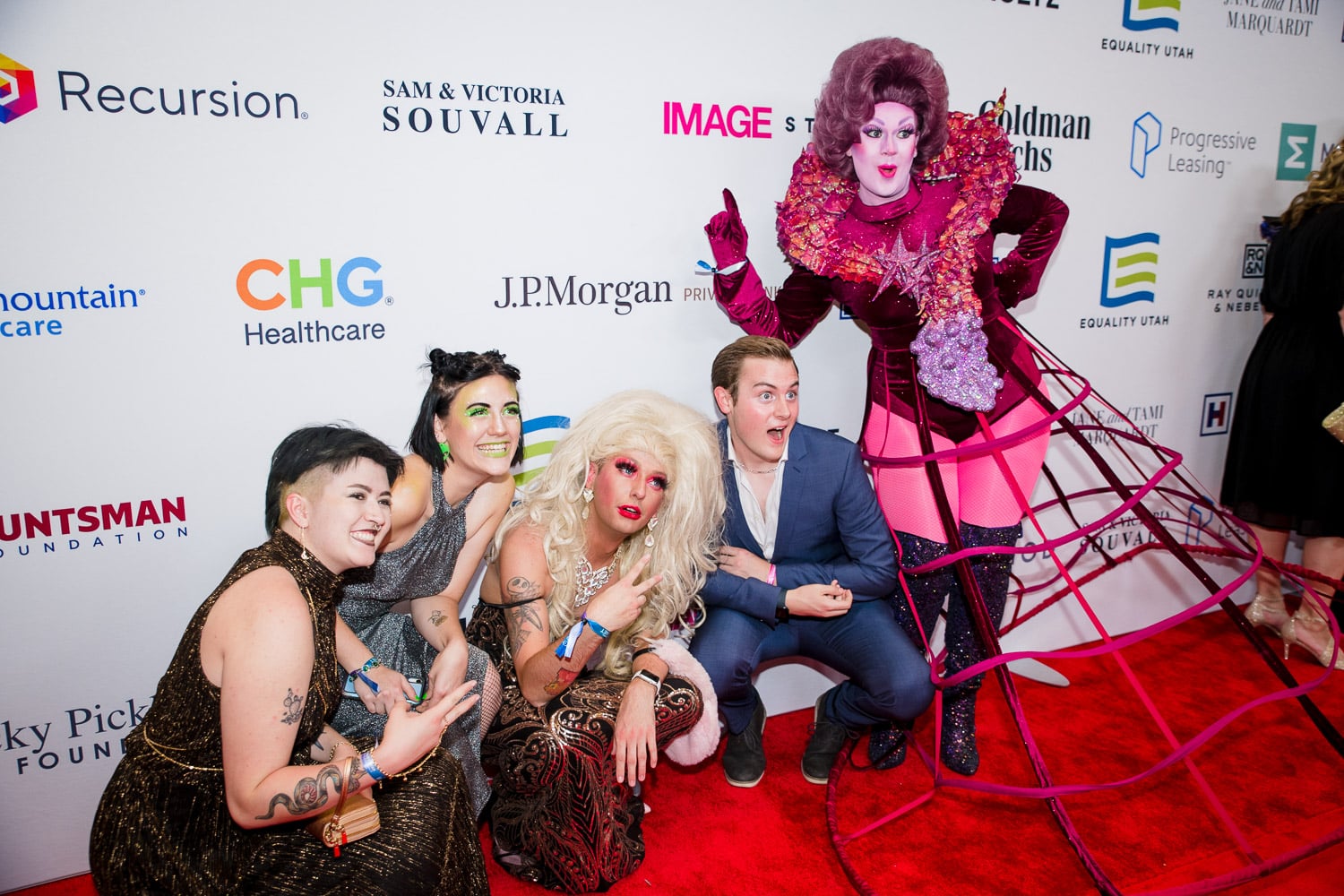 Five individuals pose together on a red carpet, some of which are dressed in drag.