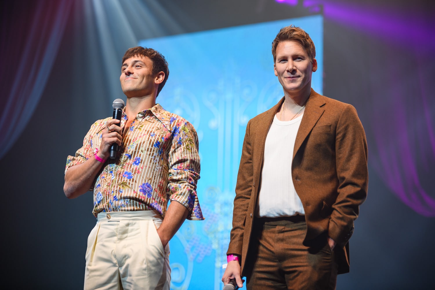 Two men stand on stage holding microphones and smiling.