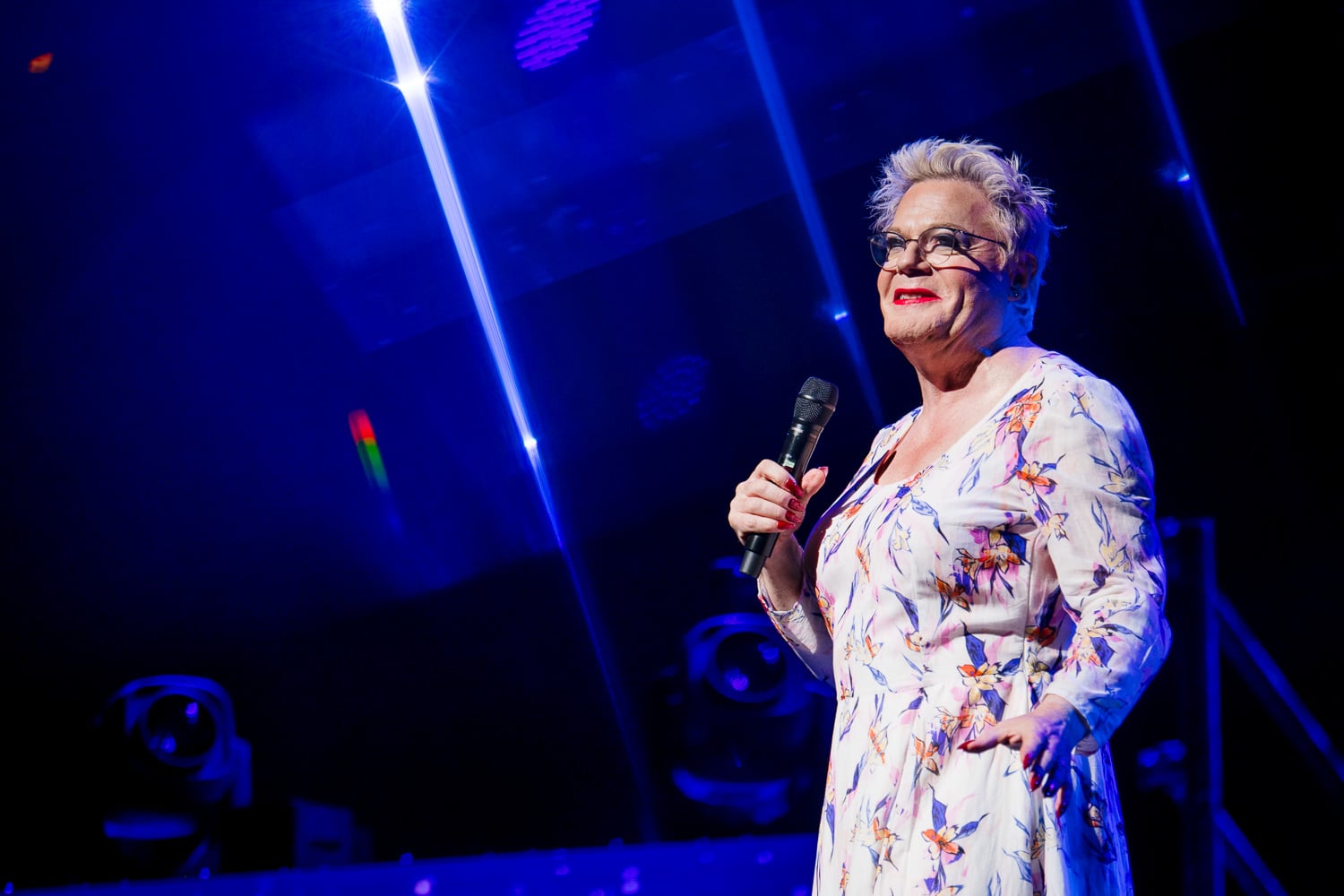 A woman in a formal dress stands on a blue stage with a microphone