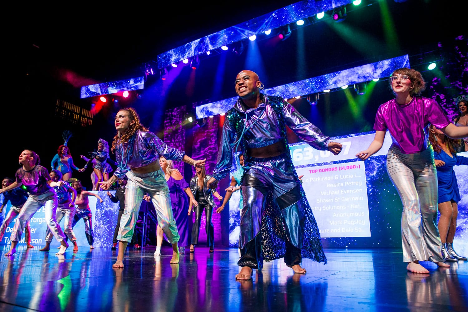 A large group of performers dance on a blue stage in shiny futuristic clothing