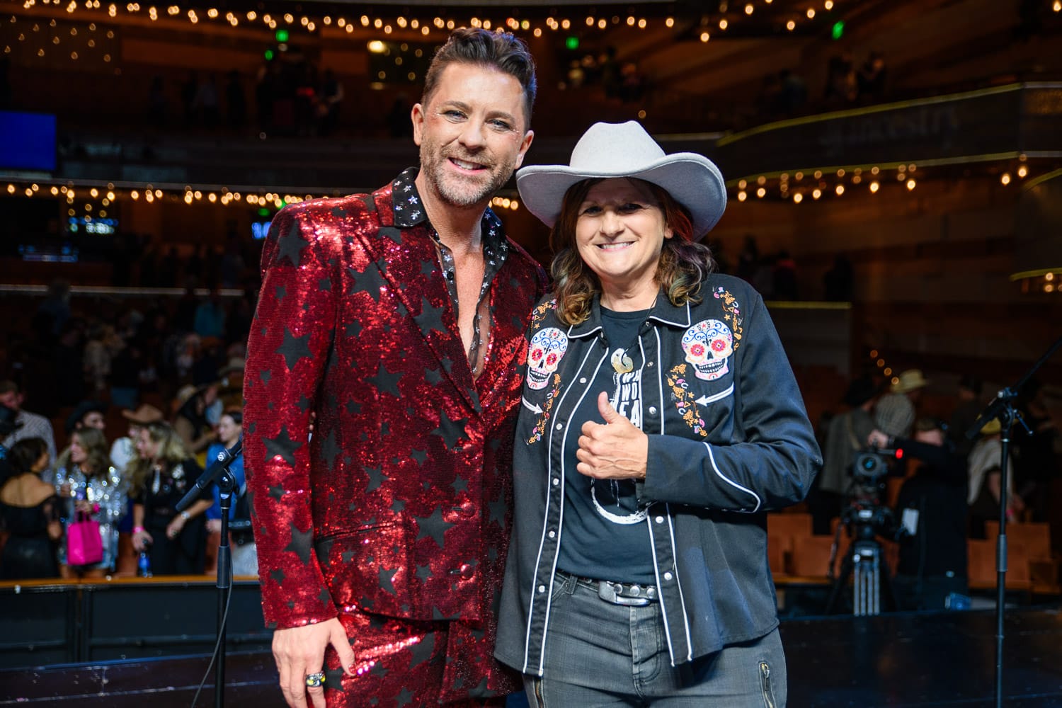 Man and woman on stage pose together. The man on the left is dressed in a shiny red suit next to a woman dressed in western attire giving a thumbs up to the camera.