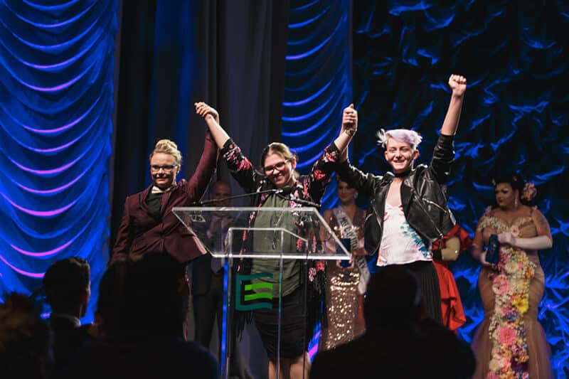 Three individuals raise their arms together on a blue stage