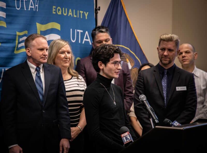 An individual stands behind a podium at a press conference with an Equality Utah backdrop behind them. There is a group of people standing with them.