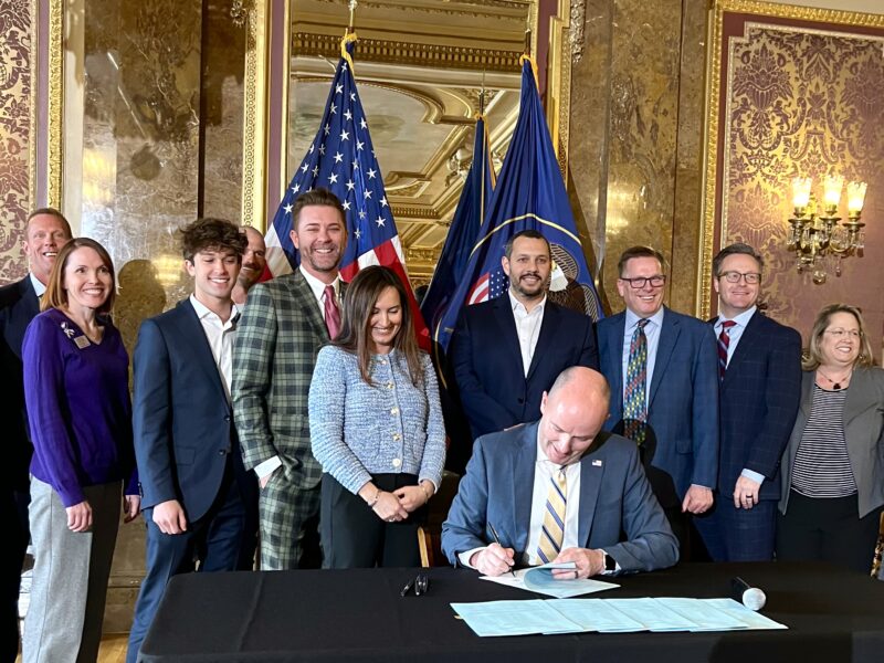 A man sits at a table signing a bill with a group of people behind him smiling