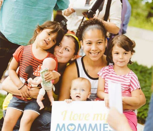 A family poses together outside, a baby holds a sign that says 'I love my Mommies'