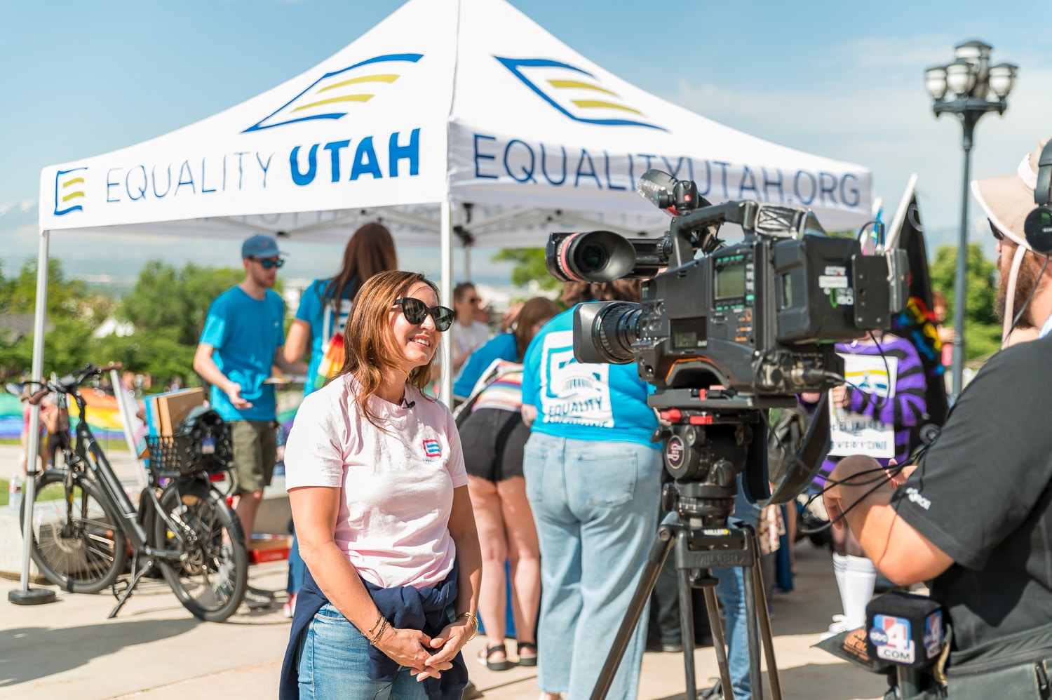 A woman stands in front of a news camera. In the background, there is a tent featuring 'EQUALITYUTAH.ORG'