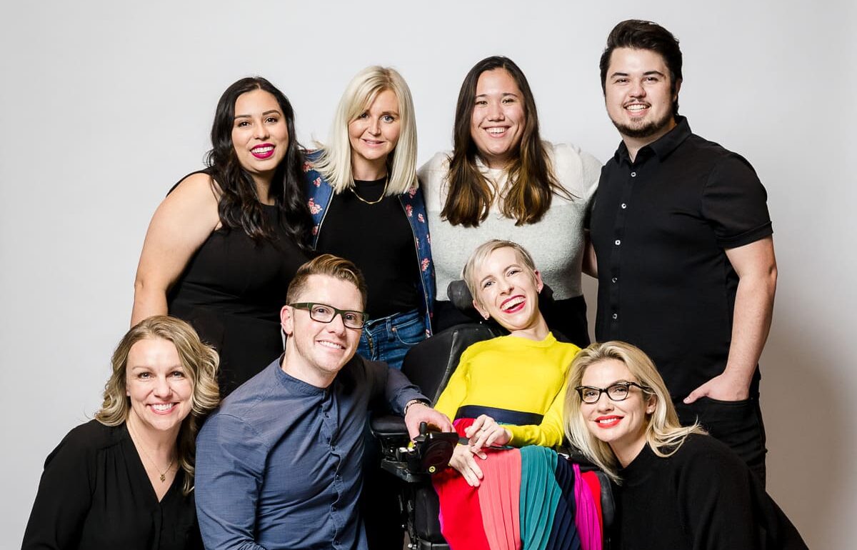 Group photo of eight people posing in front of a plain white background. The individuals are smiling and dressed in casual or semi-formal attire. A woman in a wheelchair wearing a vibrant yellow top and a colorful skirt is in the center of the group.