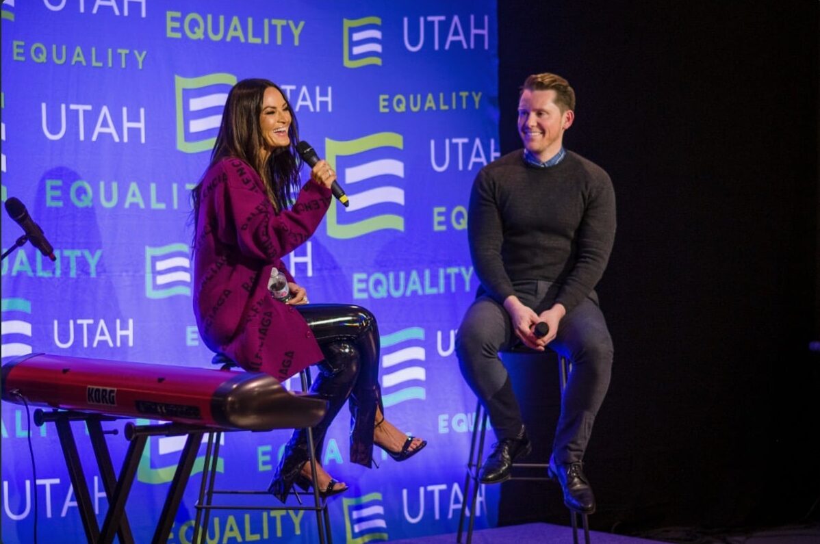 Two individuals on a stage in front of a blue backdrop with 'Equality Utah' logos. The person on the left, wearing a red patterned jacket and black shiny pants, is speaking into a microphone while seated on a stool near a keyboard. The person on the right, dressed in a dark sweater and jeans, is seated on a stool, smiling and looking at the speaker.