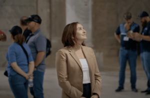 A professional photo of a woman standing indoors, wearing a beige blazer over a white top. She is looking upward with a slight smile. The background is blurred, showing other people in the casual attire.