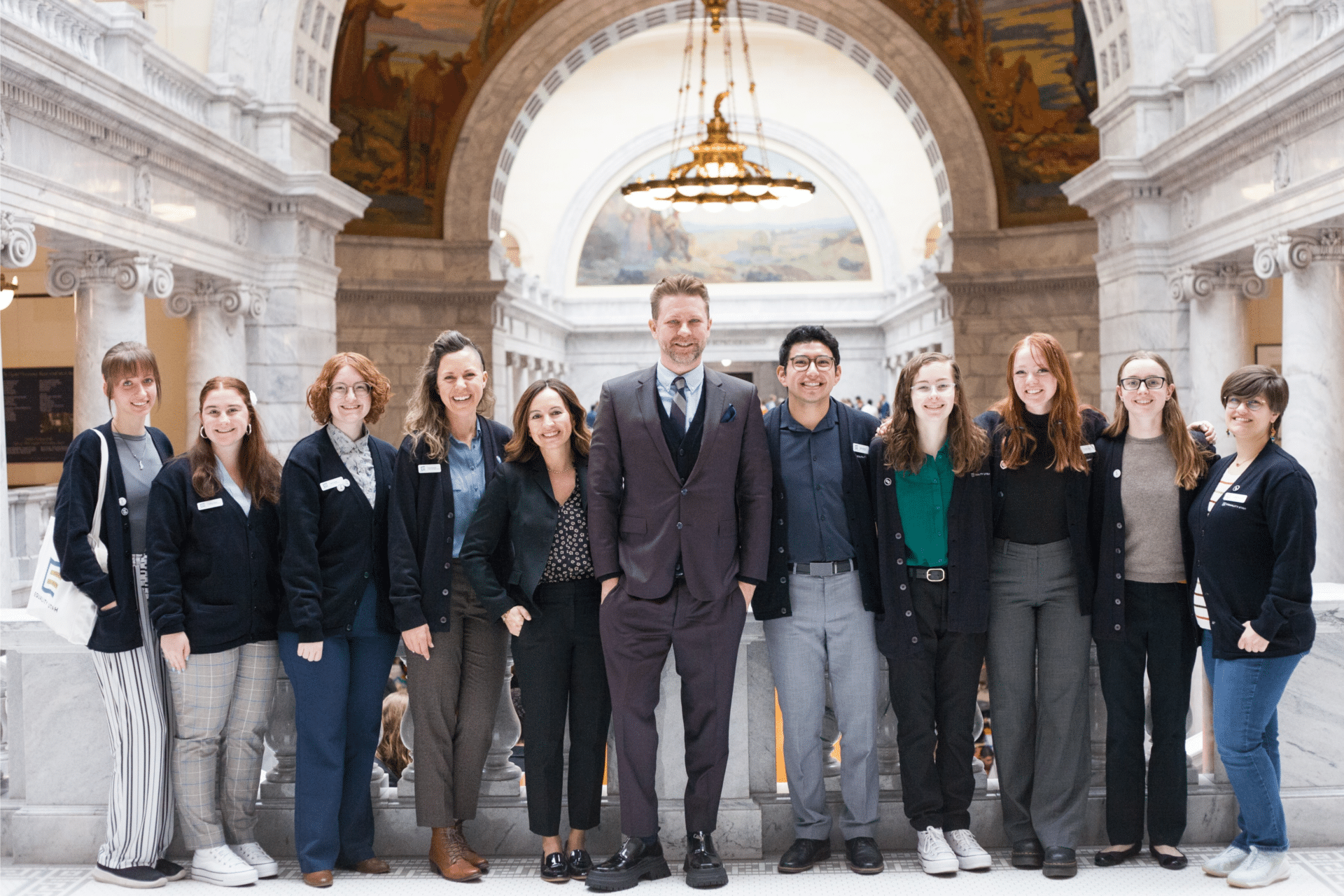 Group photo of ten people standing inside a grand building with ornate architecture, featuring arched ceilings and a chandelier. The individuals are dressed in professional attire, smiling at the camera, with Troye Williams in a suit at the center of the group.